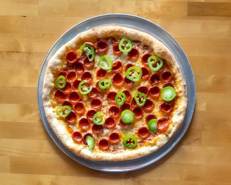 Pepperoni pizza with green pepper slices on a metal pan, on a light wooden table.