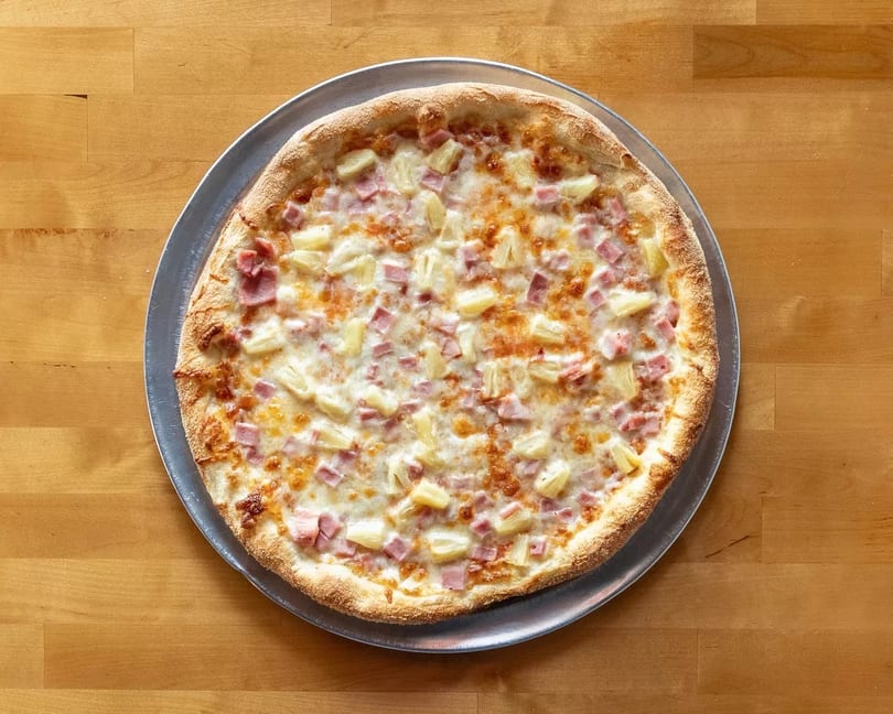 A whole Hawaiian pizza with ham and pineapple on a metal pan, on a wooden table.