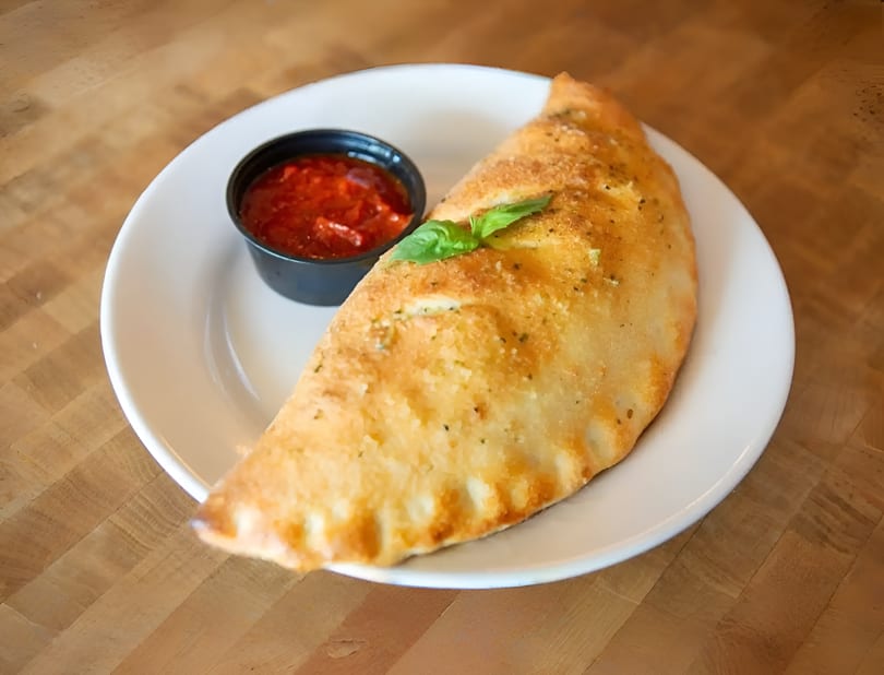 Golden-brown calzone with a basil leaf on top, served with a side of red marinara sauce.